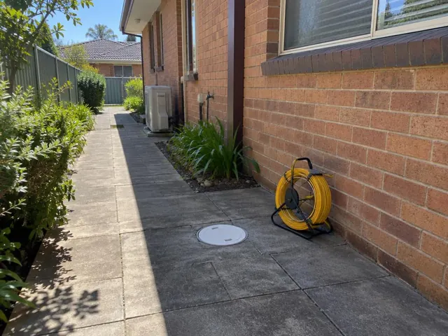 Capped drain inspection opening beside a coiled camera reel on a paved side path by a brick house in Sutherland