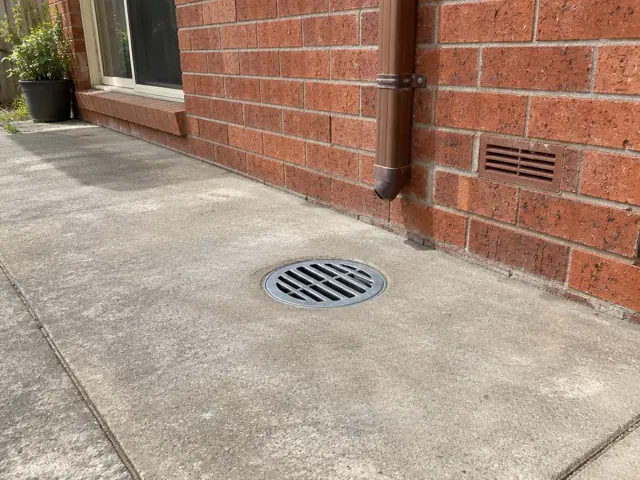 Concrete path beside a brick wall with a round metal drain grate and downpipe in Menai