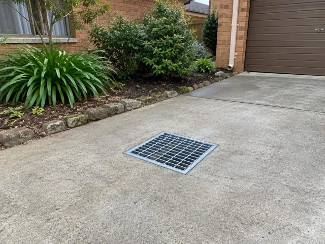 Flush stormwater grate in a concrete driveway beside a brick house and garage in Illawong